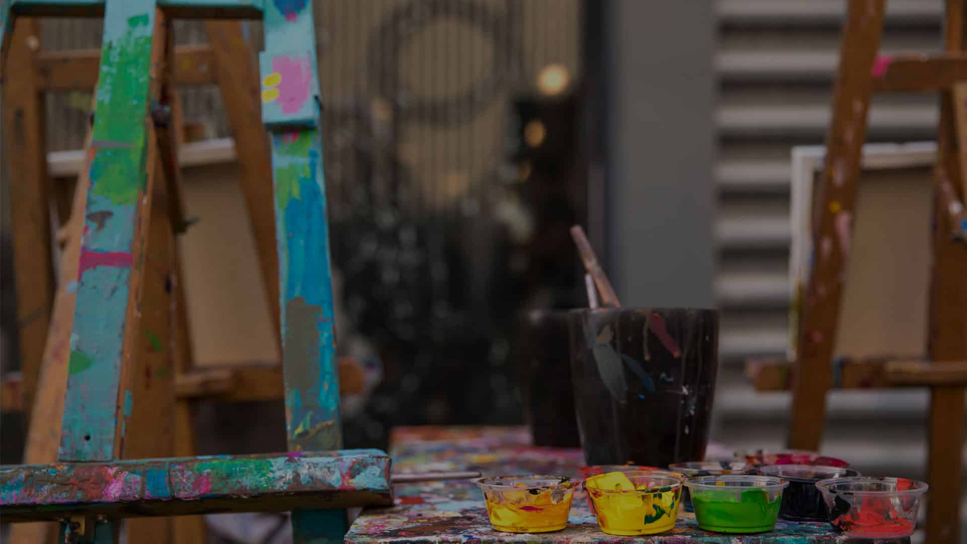 A vibrant, colorful artist's workspace featuring paint-splattered easels, a variety of paint pots filled with different colors, brushes in a dark container, and a paint-covered table surface. The backdrop resembles a studio setting rather than that of a vet clinic.