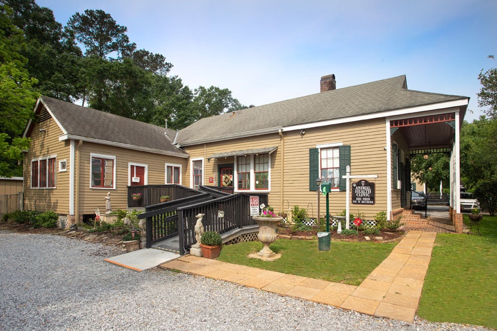 A charming, single-story, brown house with green shutters and a gable roof. The house features an accessible ramp, small potted plants, and a gravel driveway. A sign near the entrance reads “Country Store and Museum.” Trees in the background add to its appeal. It used to belong to a beloved local Vet.