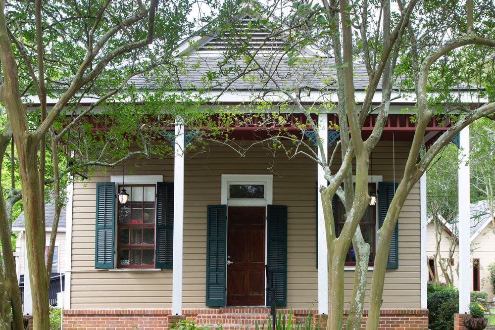 A small, single-story house with light brown wooden siding, white trim, and green shutters stands next to the local veterinarian clinic. The house has a front door with a dark finish and a small overhang. Trees and shrubs partially obscure the front, with branches extending across the view.