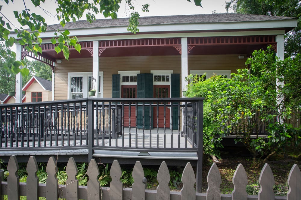 A single-story house with a large porch and a picket fence in front. The porch has dark railings and steps leading up to it. Green shutters frame the windows, and a small garden with bushes and trees surrounds the house. A neighboring vet's office is visible on the left.