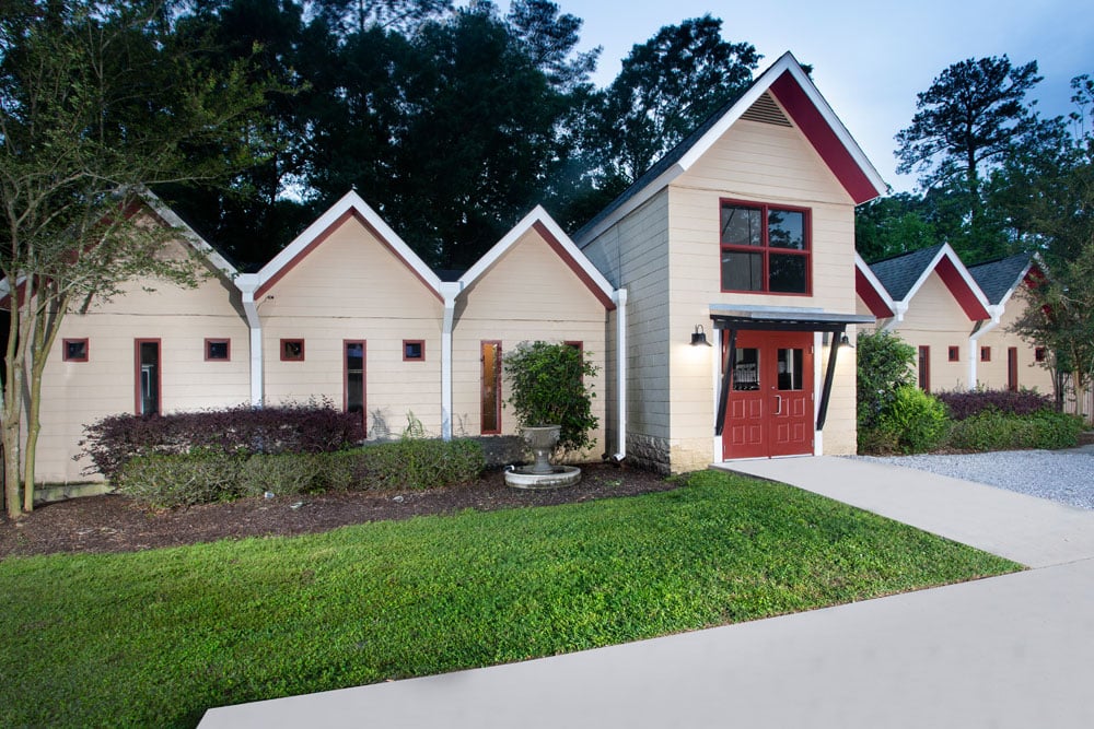 A modern single-story building with beige walls, red trim, and a series of steep gable roofs. It has several small windows and a red double-door entrance. The veterinarian clinic is surrounded by greenery, including a well-manicured lawn and bushes.