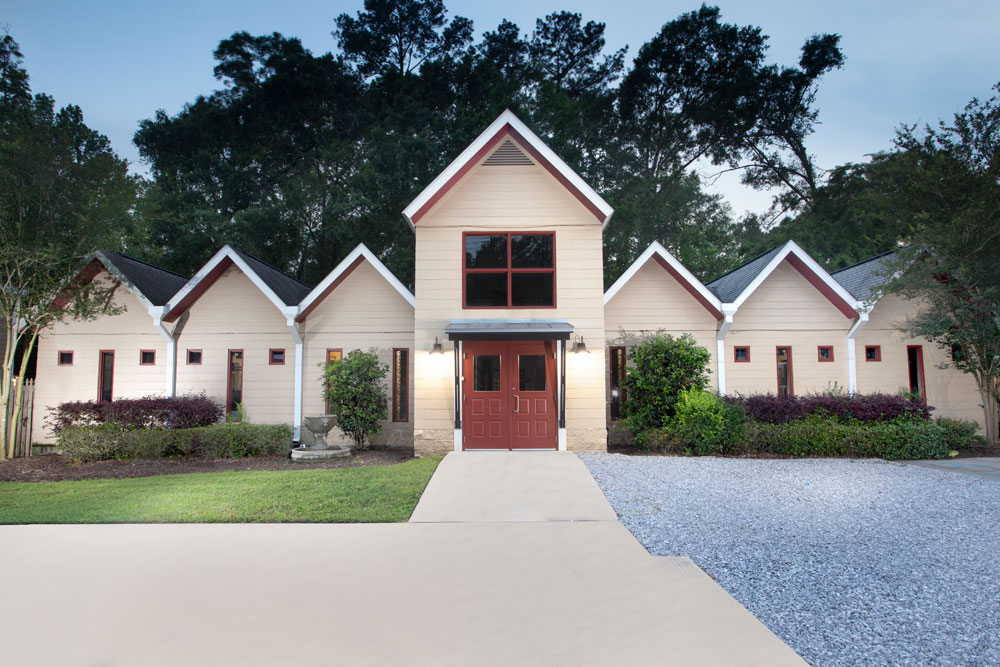 An image of a modern beige house with red double front doors, large windows, and a gravel driveway. The architecture features multiple peaked roofs and the home is surrounded by lush greenery and tall trees. The sky is overcast; it resembles the perfect retreat for a peaceful veterinarian’s home.