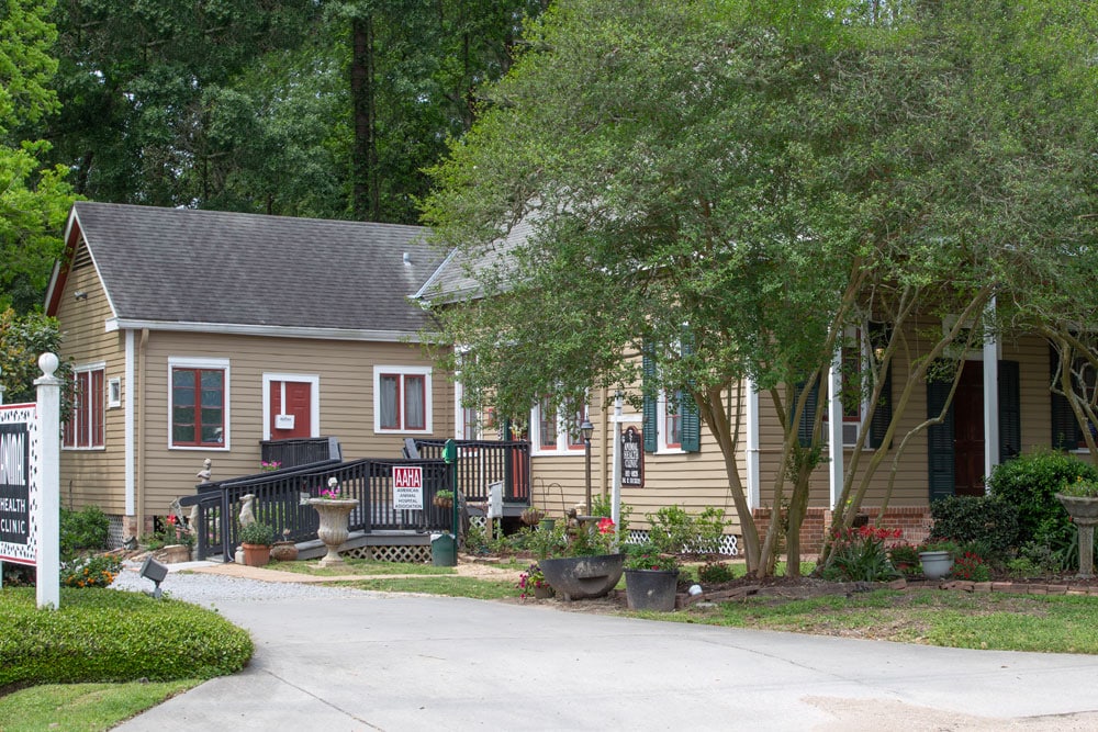 A tan-colored house with a wooden deck and black railings sits surrounded by lush trees and plants. The property features a paved driveway, potted plants, and a manicured lawn. There is a vet's for-sale sign visible on the front lawn, indicating its previous owner was an animal lover.