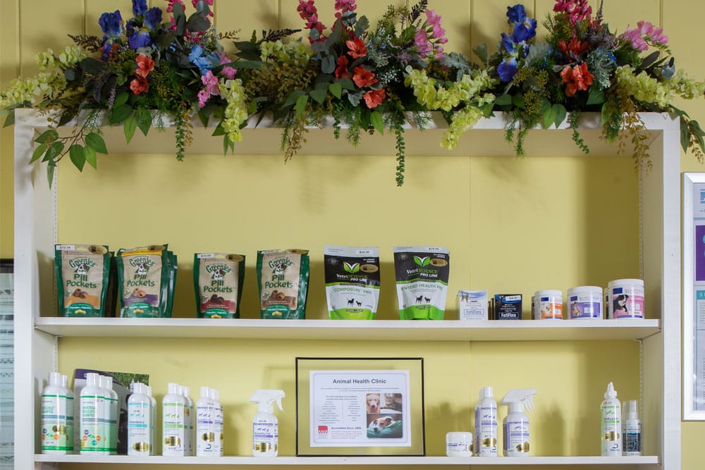 A two-tier display shelf adorned with a floral arrangement on top. The upper shelf has pet health products, including Greenies Pill Pockets and various supplements recommended by veterinarians. The lower shelf holds bottles of sprays and liquids. A framed informational sheet is centered on the bottom shelf.