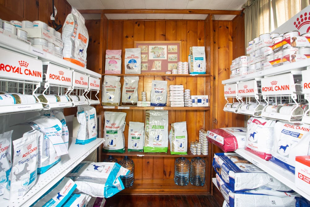 A fully stocked pet food store with wooden walls awaits you. Shelves brimming with various bags and cans of Royal Canin line each side. The bright, well-lit space also showcases water bottles below, while the back wall, decorated with pictures, adds a touch of warmth reminiscent of a veterinarian's office.