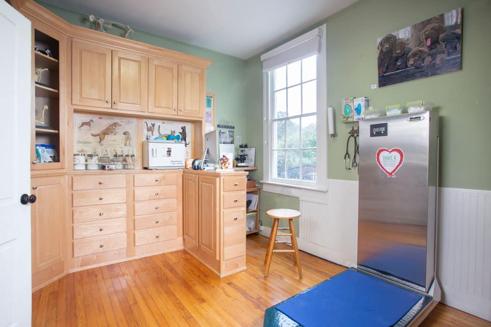 A well-organized veterinary clinic room with wooden cabinets and drawers on the left, a tall stainless steel refrigerator with a red heart sticker on the right, a scale on the floor, and a wooden stool near a window with white blinds. Various veterinarian tools and decor are visible.