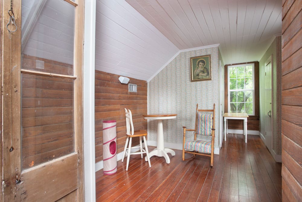 A cozy, vintage attic room with wooden walls and floors. The space features a small round table with two white chairs, a rocking chair with striped upholstery, and a framed picture of a vet on the wall. A window at the end of the room lets in natural light.