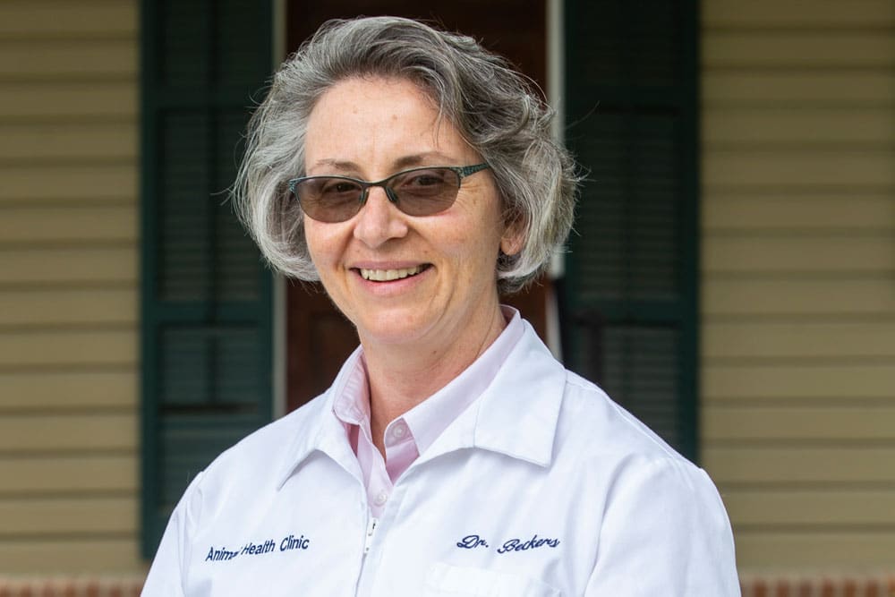 A veterinarian with short gray hair and glasses is smiling and standing in front of a beige building with green shutters. They are wearing a white coat that has "Animal Health Clinic" and "Dr. Roberts" embroidered on it.