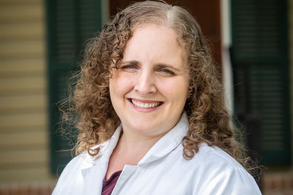 A person with curly, light brown hair smiling, wearing a white coat. The background includes a house with green shutters and a faintly visible entrance. This cheerful veterinarian exudes warmth and professionalism.